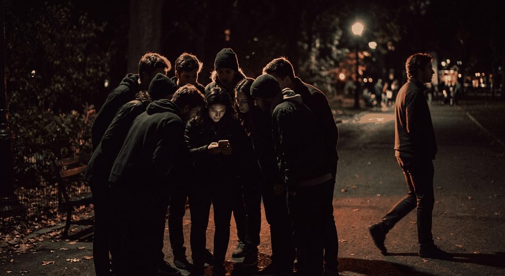 Siluet sekumpulan teman berkumpul di sekitar layar ponsel yang menyala di malam hari